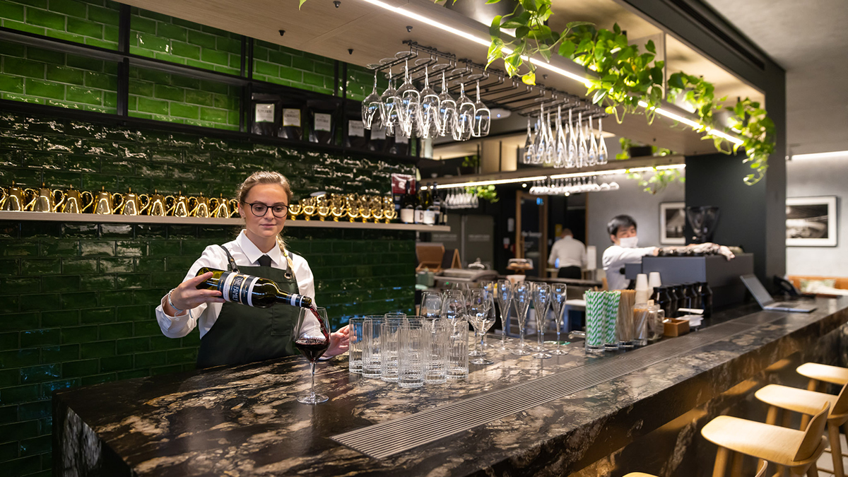 a female bartender pours a glass of red wine at a bar against a green tile background