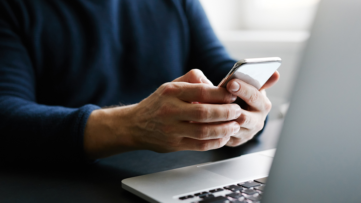 Close up of a person's hands holding a mobile phone while sitting at a laptop