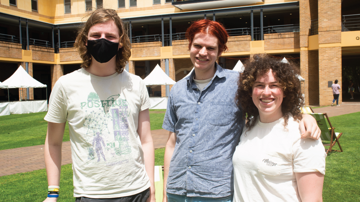 Three students stand casually together on grass on campus