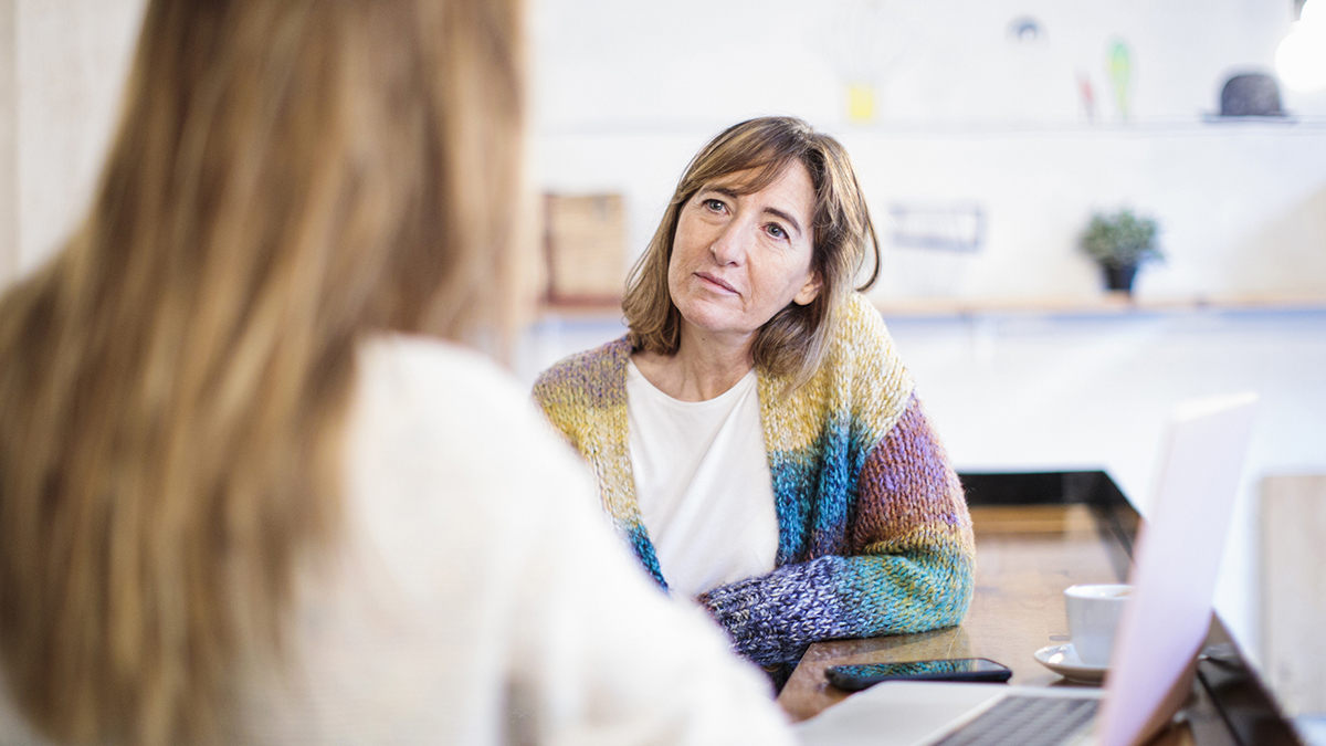 A woman in a colourful cardigan with a concerned expressions faces another woman whose back is turned