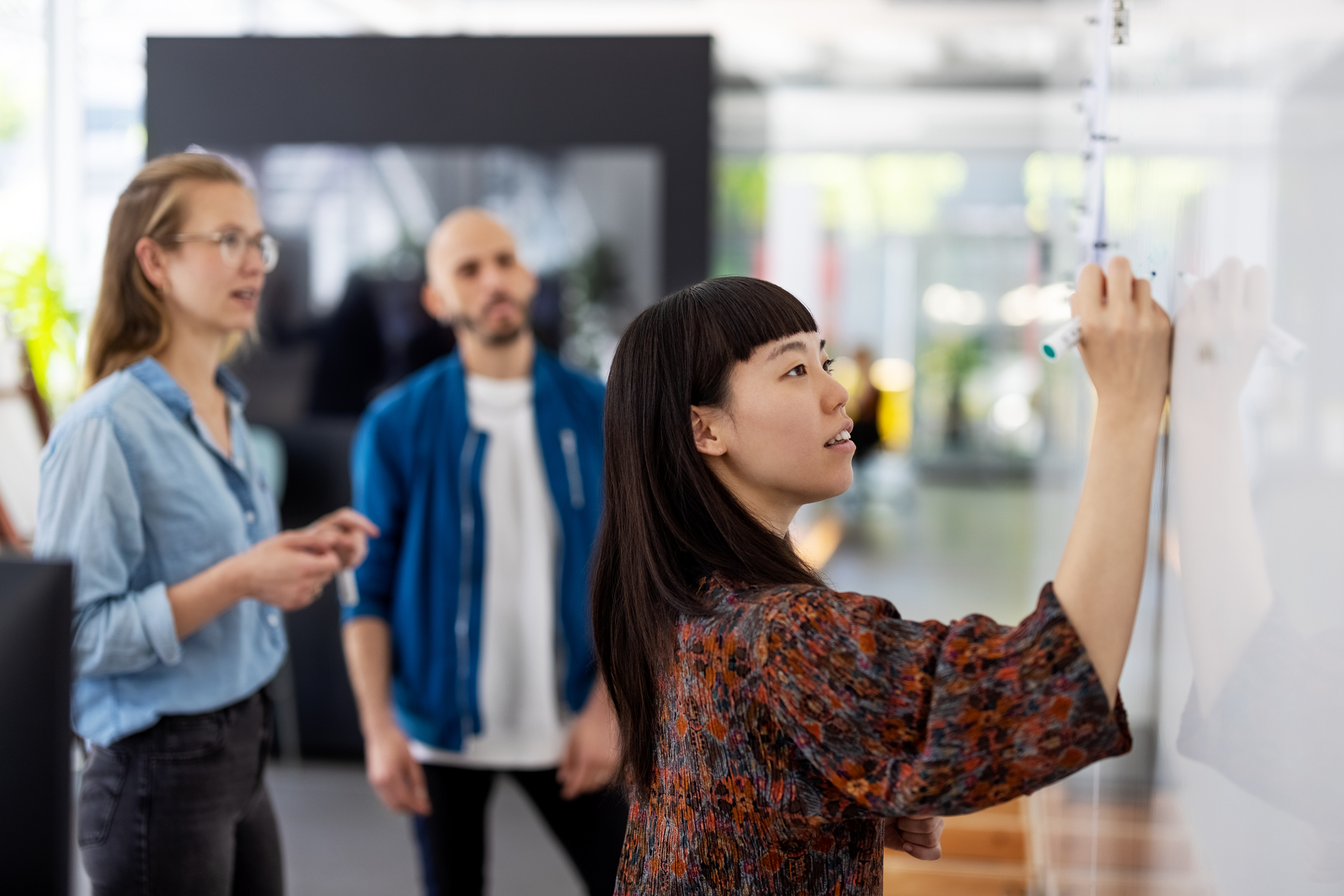 A young woman of Asian descent writes on a whiteboard in front of a male and female colleague.