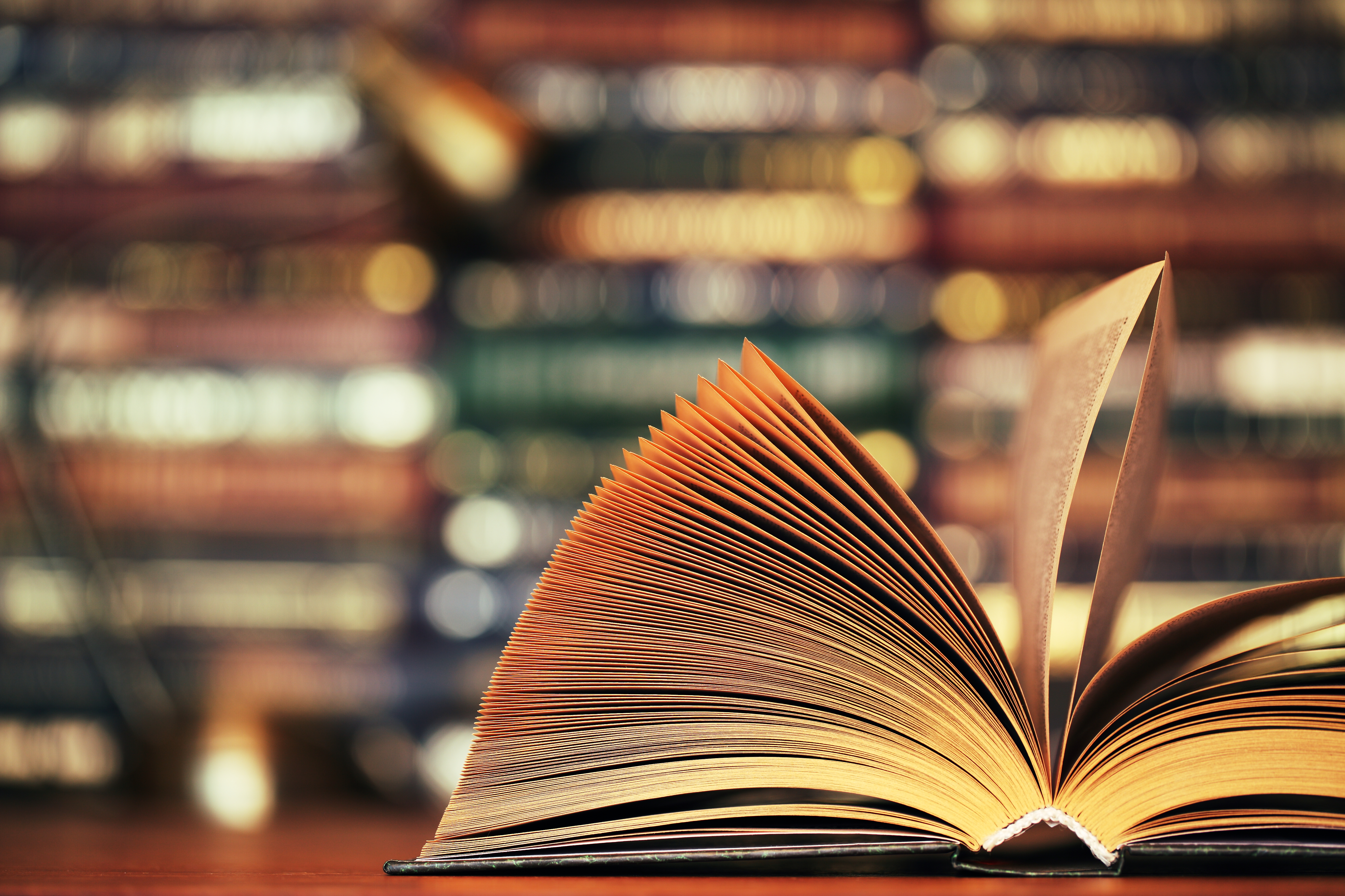 A book lies open on a table in front of a wall of books.