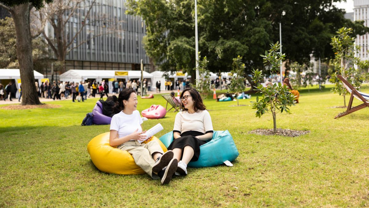 Two women sit laughing together on colourful beanbags on the lawn in front of a building