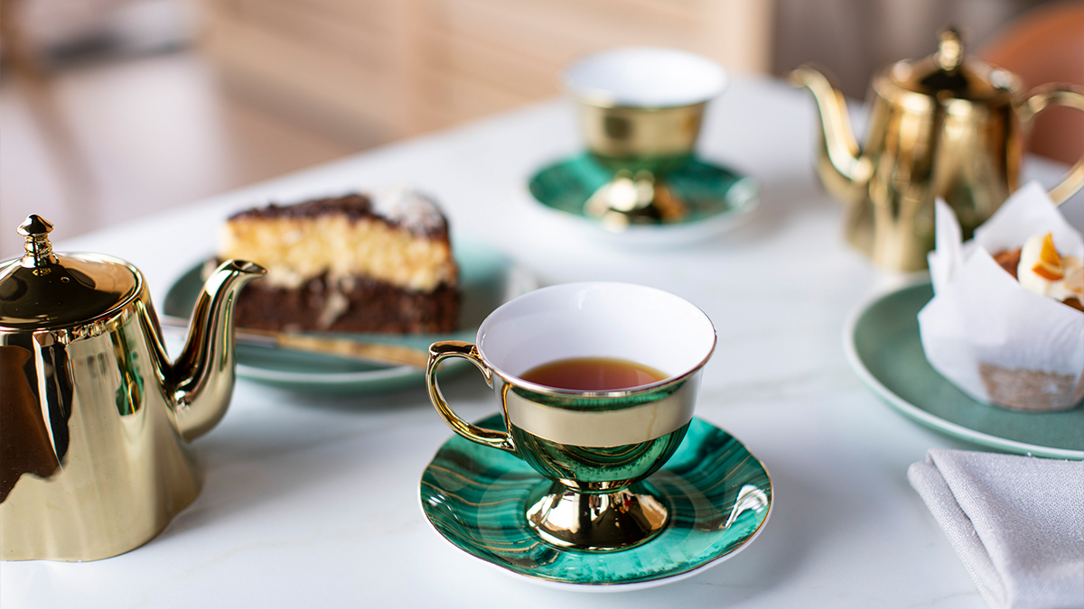 Green tea cup with black tea, a plate with cake, and a golden tea pot on a table