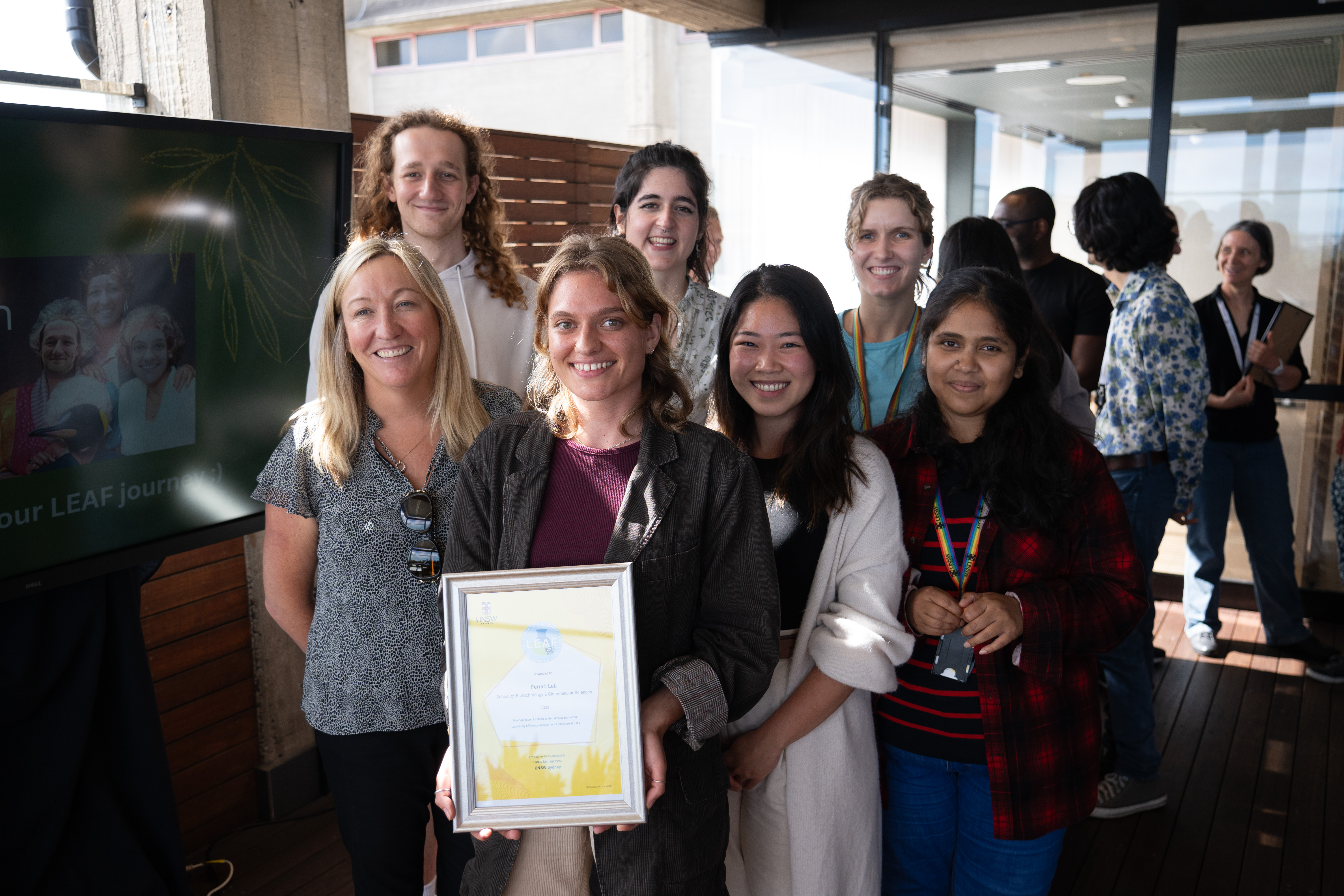 A group of seven scientists smile in front of a building, holding a framed award