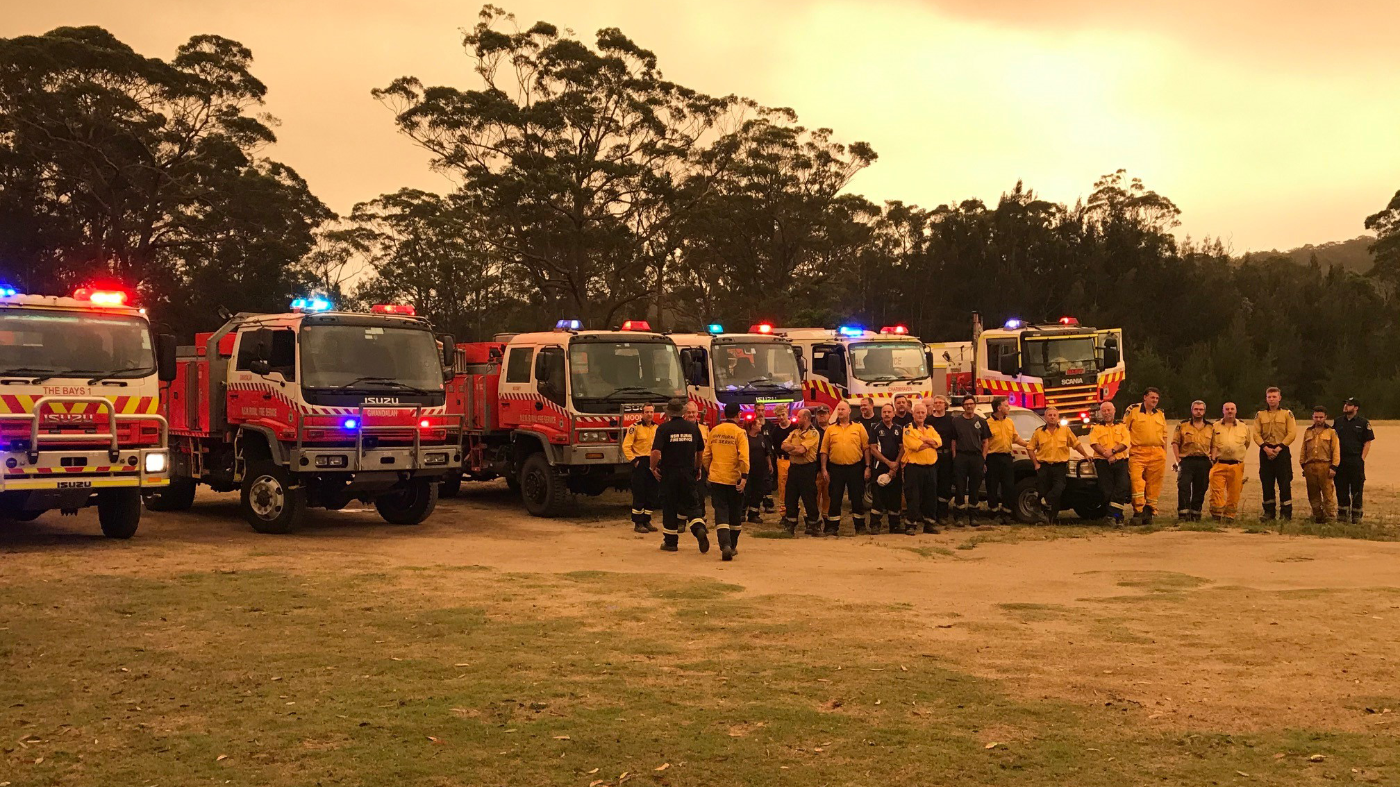 Fire trucks lined up in an open green lawn with firefighters standing in front