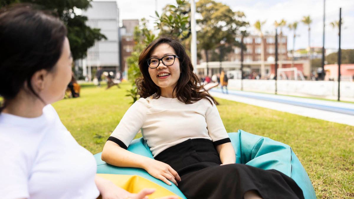 A woman wearing glasses sits on a beanbag talking to another woman outdoors.
