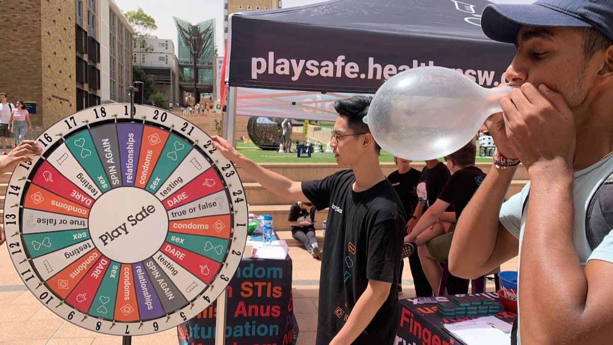 A man in a baseball cap blows up a condom at a sexual health stall with a 'Play Safe' spinning wheel