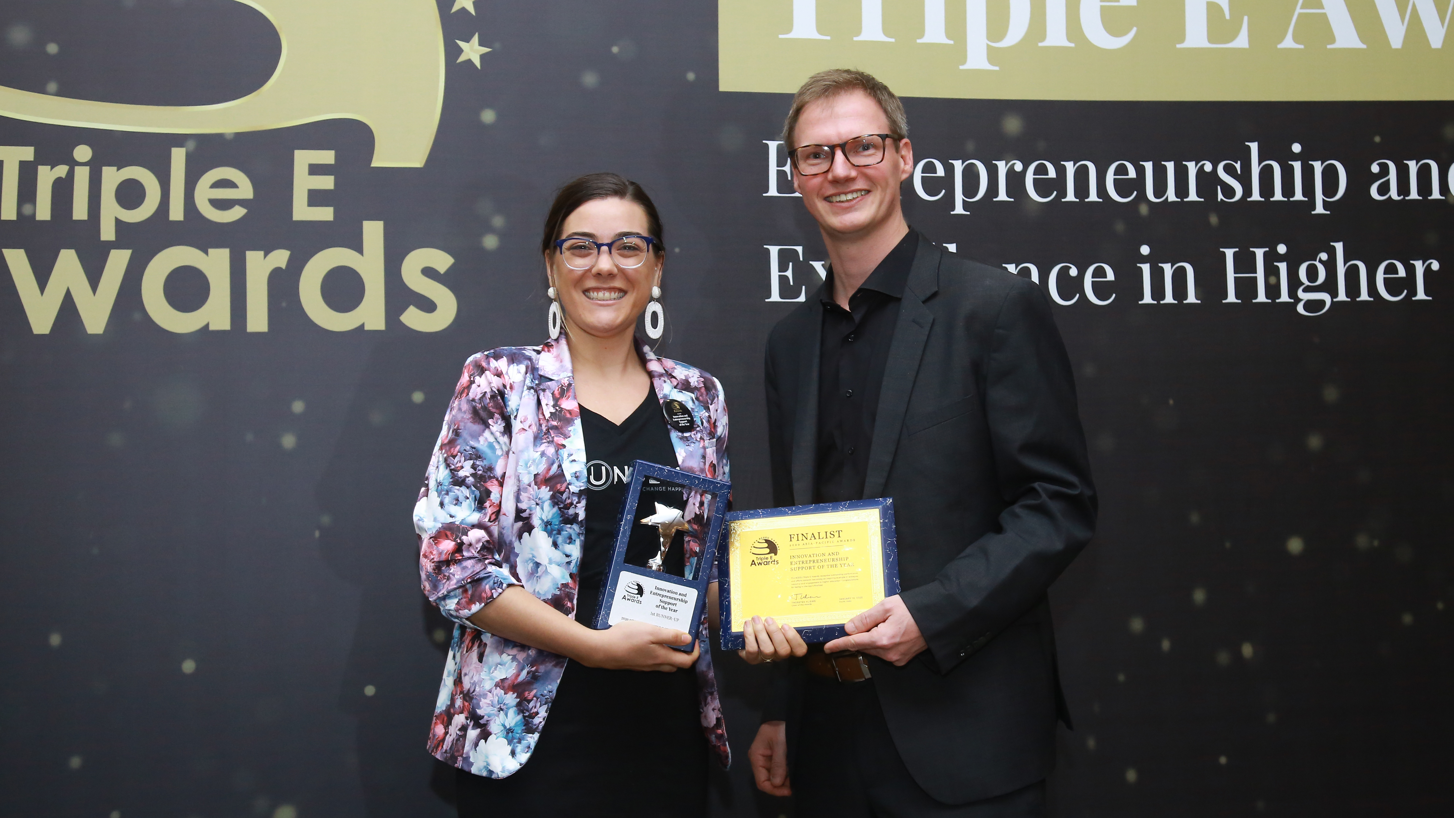 A man and woman holding an award in front of awards backdrop