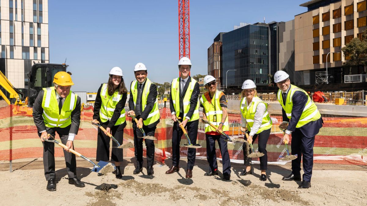 Seven men and women wearing hard hats and hi-res vests shovel earth from the ground of a construction site