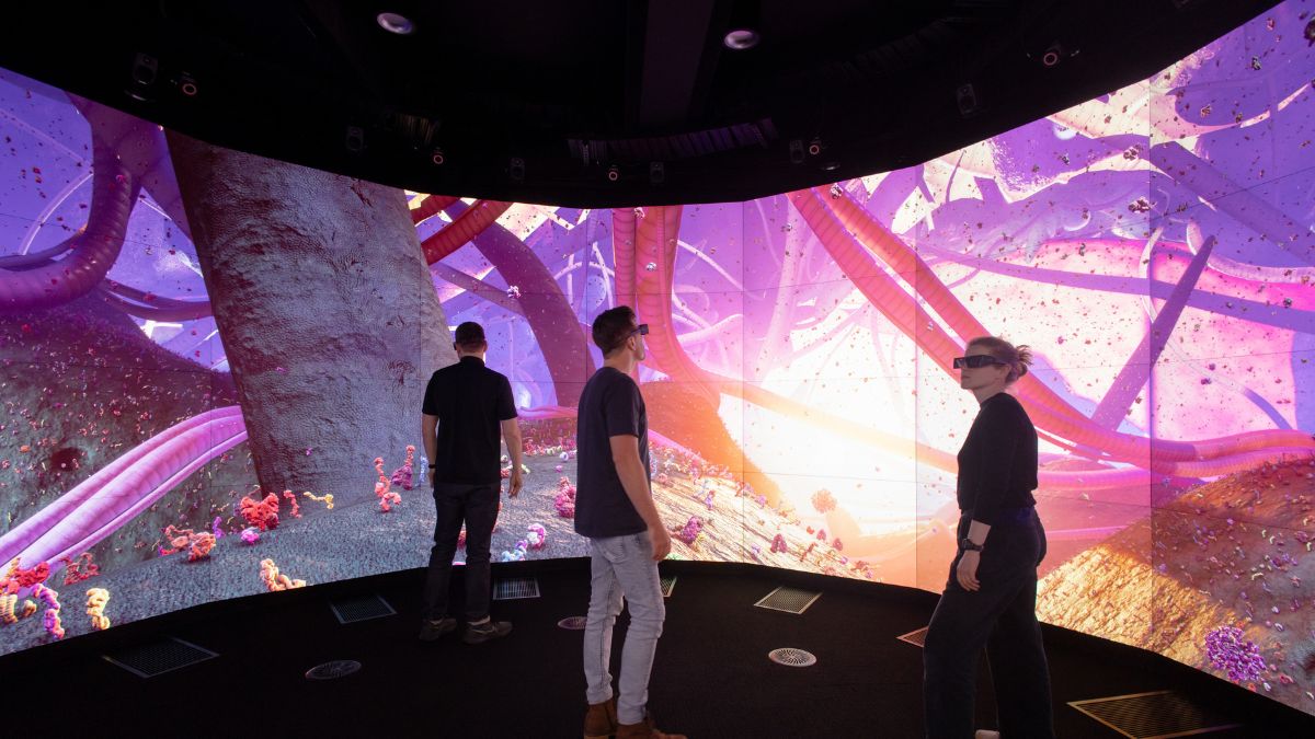 A blonde woman wearing a virtual reality headset and two men stand in front of a colourful circular life-size screen