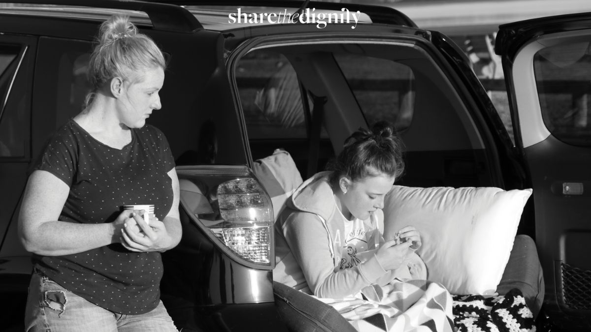 A woman stands beside the open boot of a car where a girl sits with a pillow and blanket