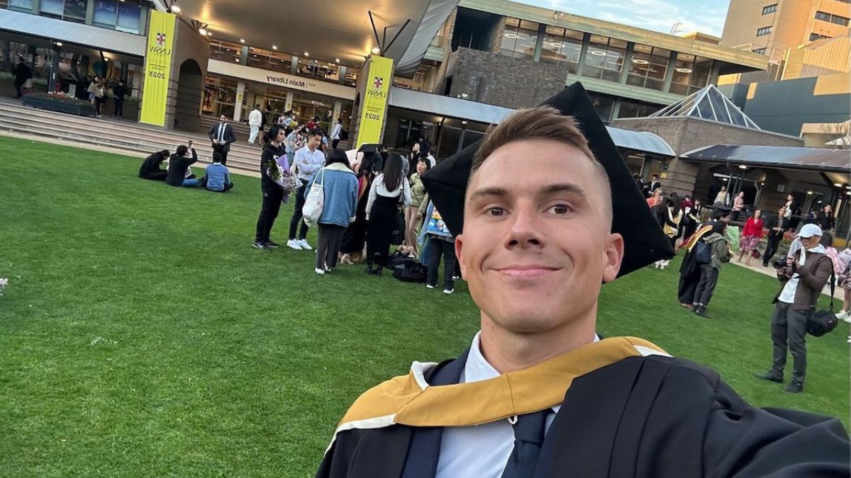 A male graduate in black cap and gown with a yellow hood stands on the lawn before a building