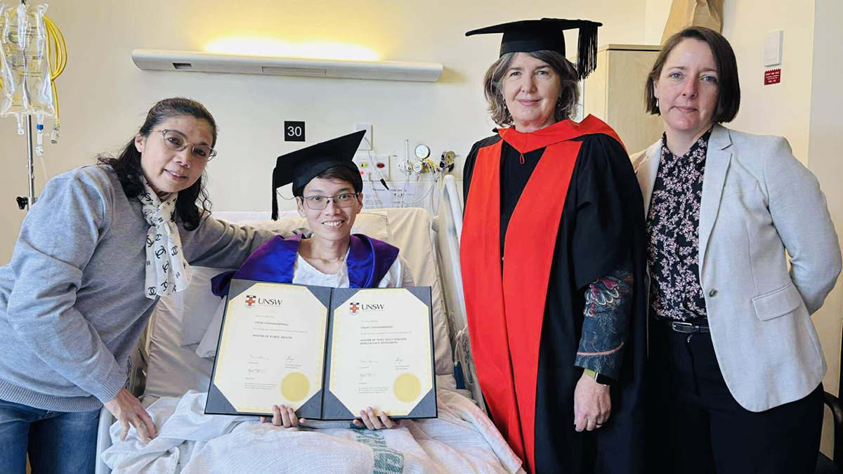 Vinay Chansomphou, a man wearing a graduation cap, in a hospital bed with three women standing at the bedside, Vinay's mother, Vinay, Rebecca Ivers and Anita Heywood (L-R)