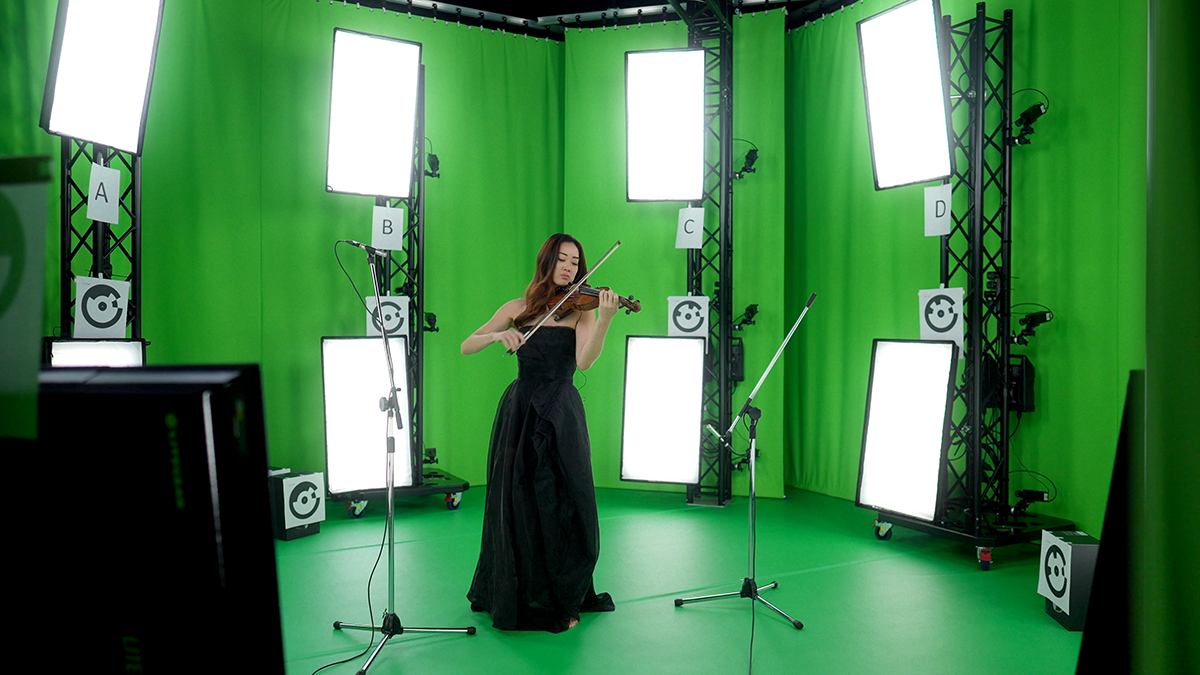 A woman in a long black dress playing violin in a green screen studio with multiple cameras and lights
