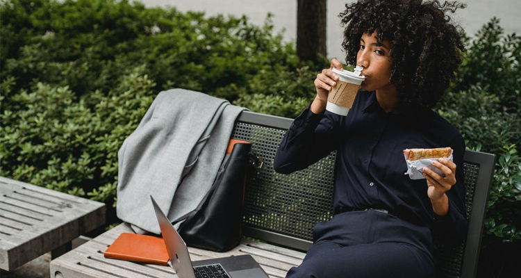 Woman eating and drinking with laptop on bench