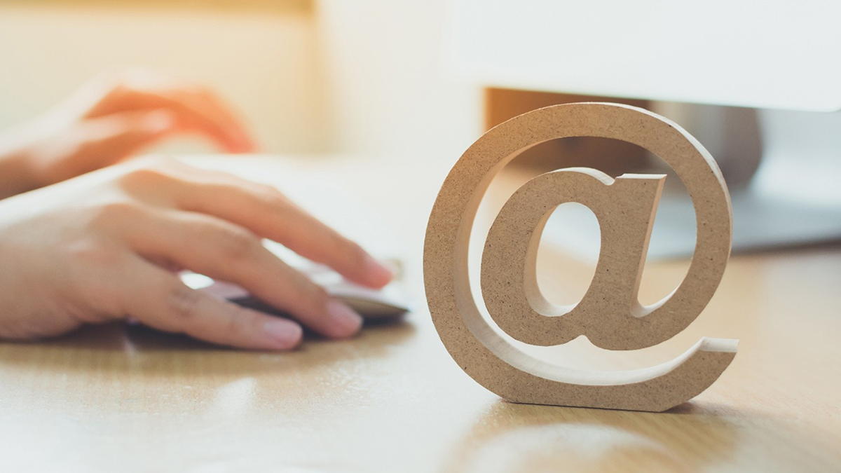 A wooden @ symbol on a table with a person's hands using a mouse in the background