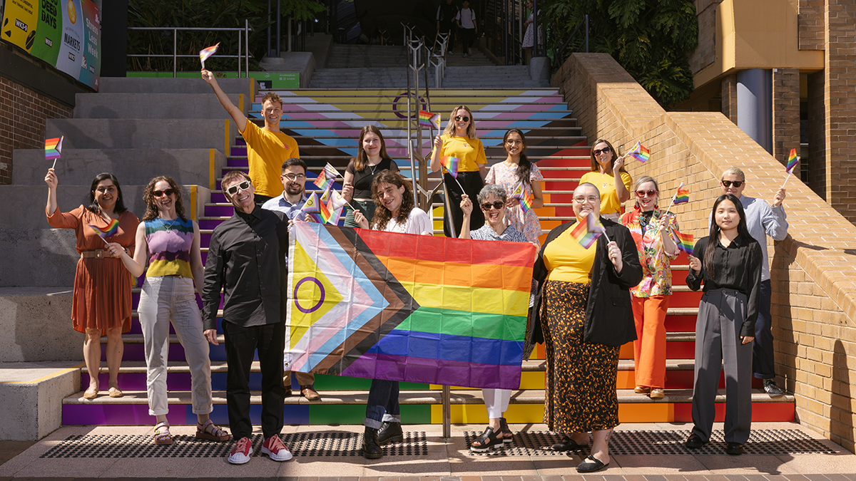 A group of people gathered on the Basser steps with a Progress Pride flag