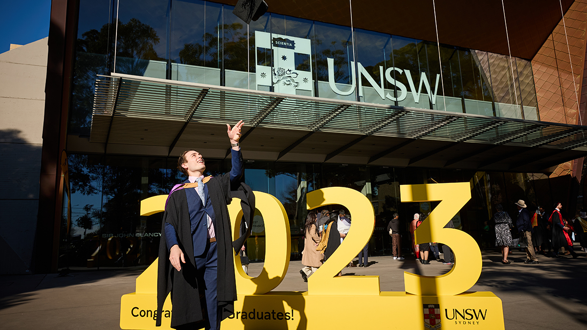 A man in graduation regalia standing in front of a sign that says 2023 throw his graduation cap in the air