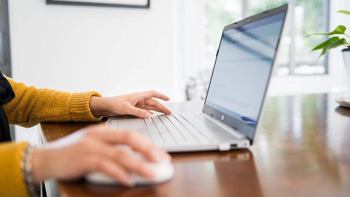 Close up of a laptop on a wooden table and a woman in a mustard sweater with her hands on the mouse