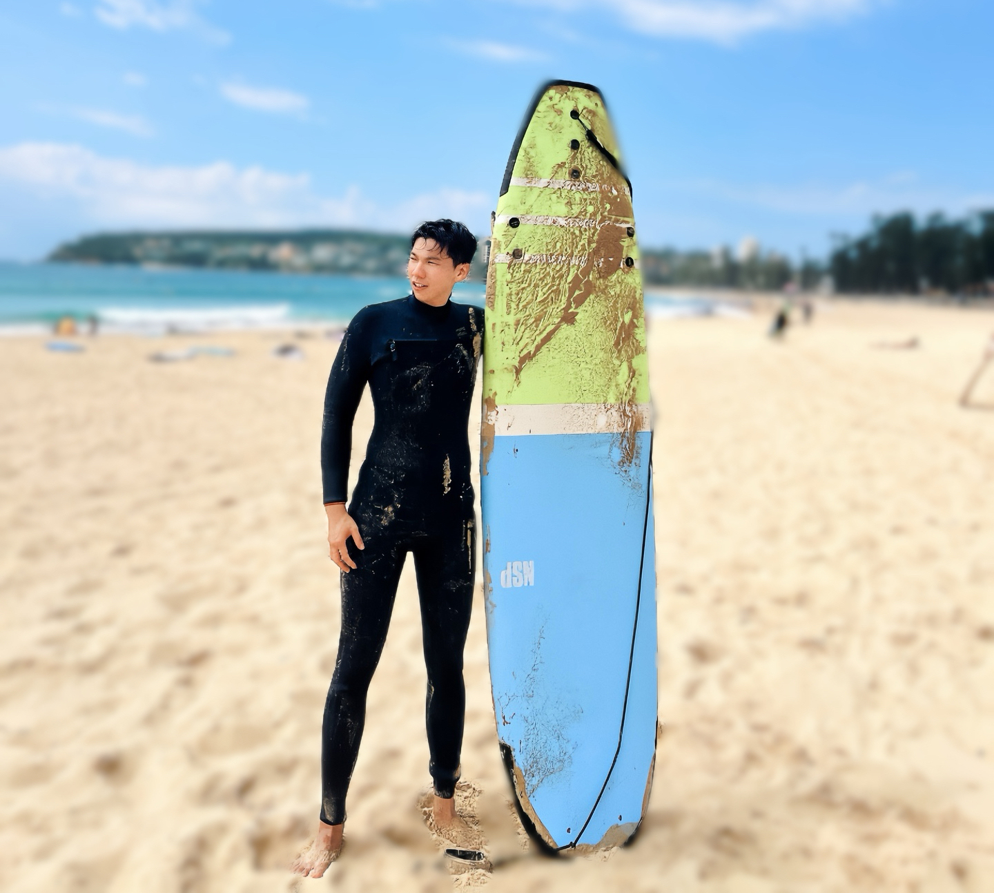 A young man in black wetsuit standing on the beach with a blue and yellow longboard