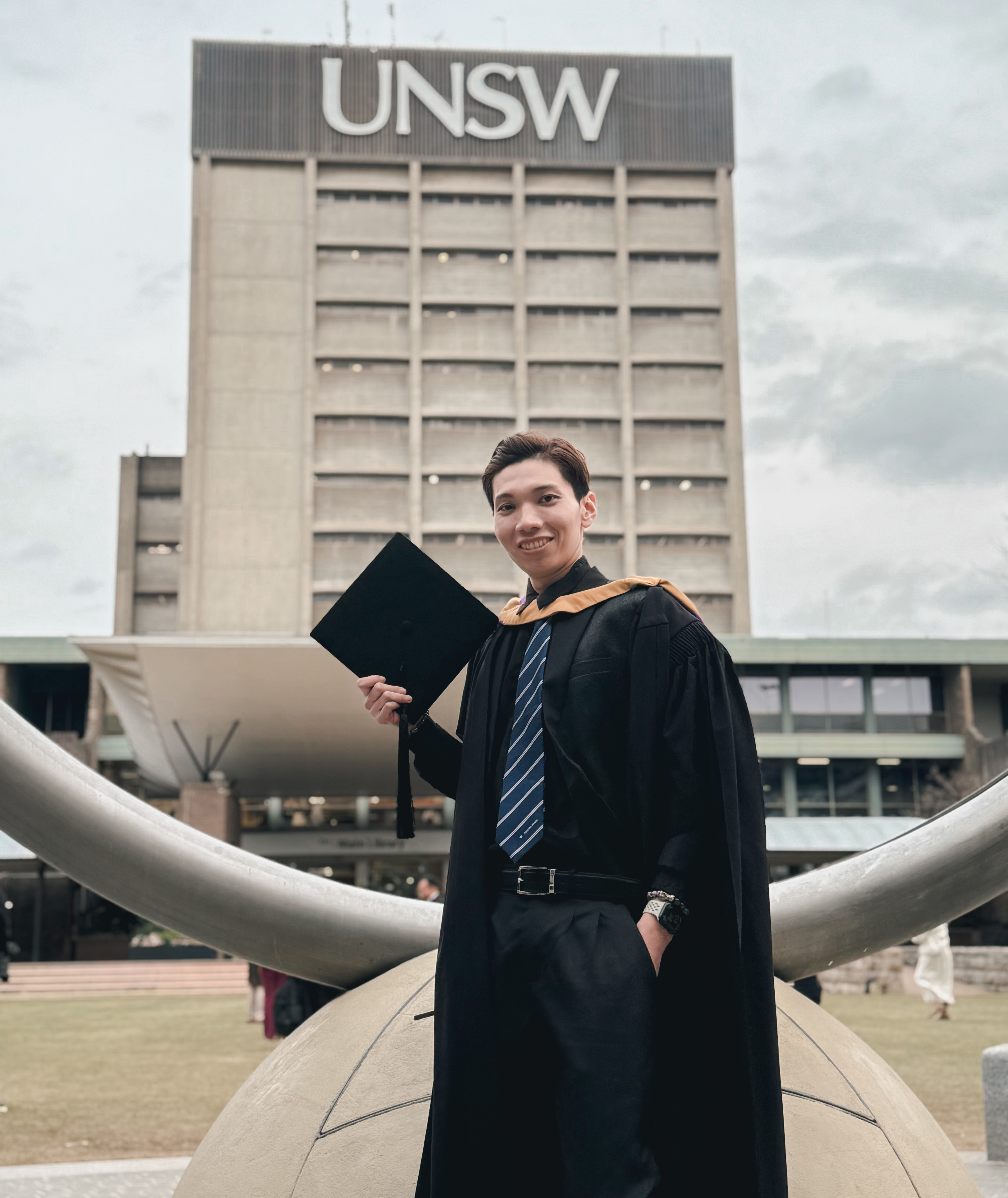 A young man in graduation gown holding a graduation cap in front of the UNSW library tower