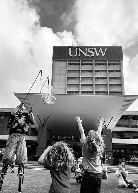 Two children, backs turned, watch a balloon artist on stilts on the lawn in front of a building