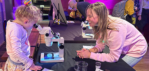 A woman smiling at a table where a young girl looks through a microscope