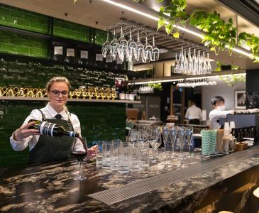 a female bartender pours a glass of red wine at a bar against a green tile background