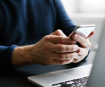 Close up of a person's hands holding a mobile phone while sitting at a laptop