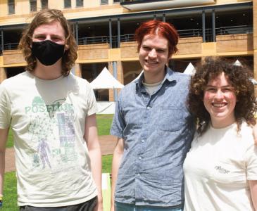 Three students stand casually together on grass on campus