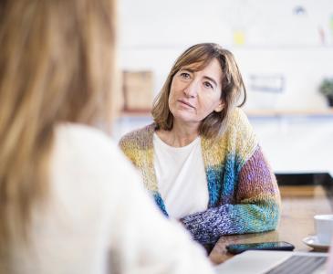 A woman in a colourful cardigan with a concerned expressions faces another woman whose back is turned