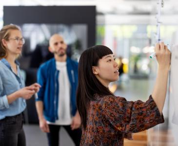 A young woman of Asian descent writes on a whiteboard in front of a male and female colleague.