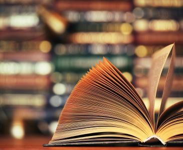 A book lies open on a table in front of a wall of books.