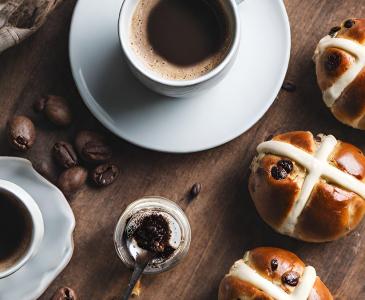 Hot cross buns on a wooden bench beside white cup with coffee