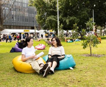 Two women sit laughing together on colourful beanbags on the lawn in front of a building