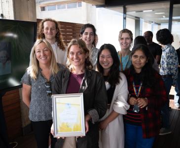 A group of seven scientists smile in front of a building, holding a framed award