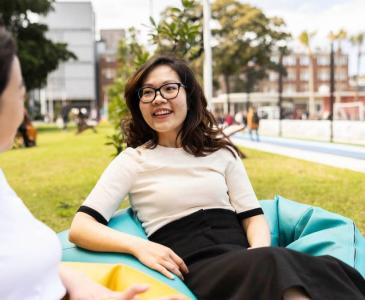 A woman wearing glasses sits on a beanbag talking to another woman outdoors.