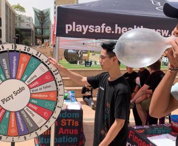 A man in a baseball cap blows up a condom at a sexual health stall with a 'Play Safe' spinning wheel