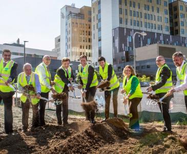 A group of men and women in hi-vis vests shovel dirt on an urban construction site.