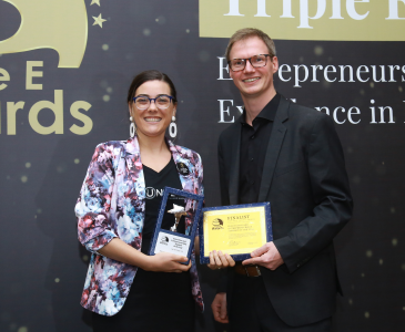 A man and woman holding an award in front of awards backdrop