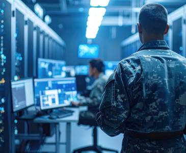 A man in an army uniform walking into a data centre with another man working on a computer.