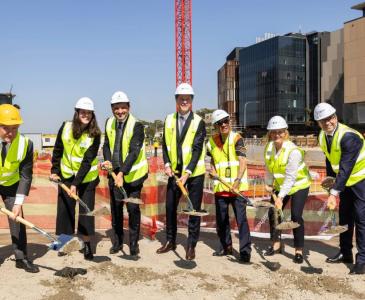 Seven men and women wearing hard hats and hi-res vests shovel earth from the ground of a construction site