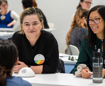 Two female staff members sit at a table in a room full of people