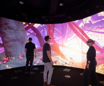 A blonde woman wearing a virtual reality headset and two men stand in front of a colourful circular life-size screen
