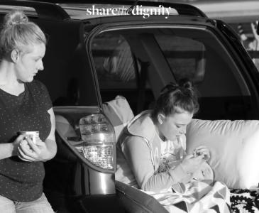 A woman stands beside the open boot of a car where a girl sits with a pillow and blanket