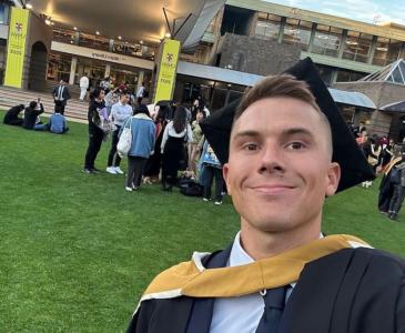 A male graduate in black cap and gown with a yellow hood stands on the lawn before a building