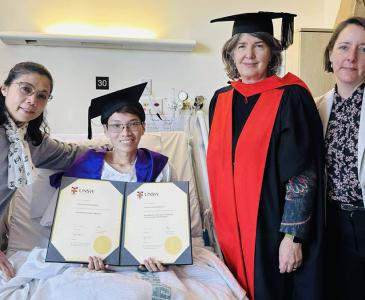 Vinay Chansomphou, a man wearing a graduation cap, in a hospital bed with three women standing at the bedside, Vinay's mother, Vinay, Rebecca Ivers and Anita Heywood (L-R)