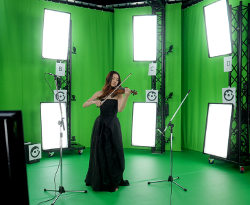 A woman in a long black dress playing violin in a green screen studio with multiple cameras and lights