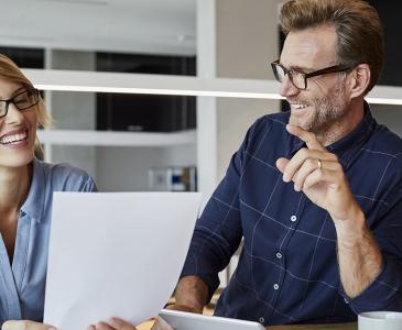 A woman with blonde hair holds a document while seated beside a man with brown hair